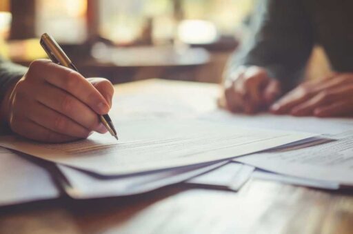 Close-up of a hand signing paperwork on a table with another person nearby