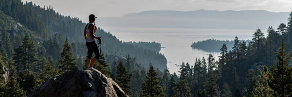 Person standing on a rock, overlooking a pine-covered valley and large lake