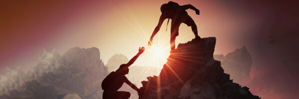 Silhouette of one person helping another climb a mountain peak at sunrise